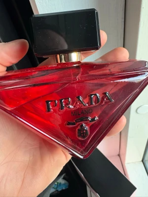 Paradoxe - Snygg parfym från Prada i en djup röd, triangelformad glasflaska med svart och silverdetaljer. Flaskan har en stor svart fyrkantig kork och tydlig Prada-logga på framsidan. Perfekt för dig som gillar exklusiva dofter och vill ha en statement-flaska på hyllan.