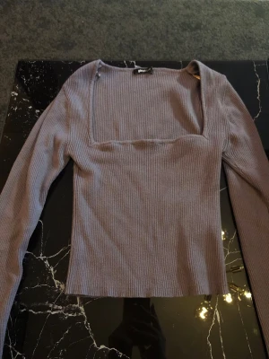 Ljusgrå/lila ribbad topp Gina Tricot - Trendig ljuslila ribbad topp från Gina Tricot med lång ärm och fyrkantig urringning och lite volang vid ärmen. Toppen är croppad och har en slim passform som sitter snyggt på kroppen. Perfekt att styla med höga jeans eller kjol.