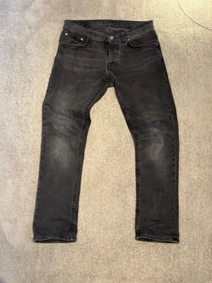 Svarta slim fit jeans från Nudie Jeans - Säljer ett par svarta slim fit jeans från Nudie Jeans med snyggt slitna detaljer och klassisk femficksmodell. Jeansen har en diskret tvättad look och raka ben. Perfekta för dig som gillar en stilren och avslappnad vibe.