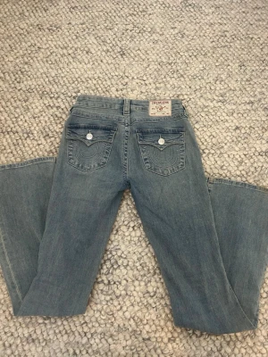 Blå bootcut jeans från True Religion - Säljer ett par blå bootcut jeans från True Religion med klassiska kontrastsömmar och snygga fickdetaljer bak. Jeansen har knappar och nitar i silverfärg samt en patch med logga bak i midjan. Tillverkade i mjuk denim med lätt tvättad look.