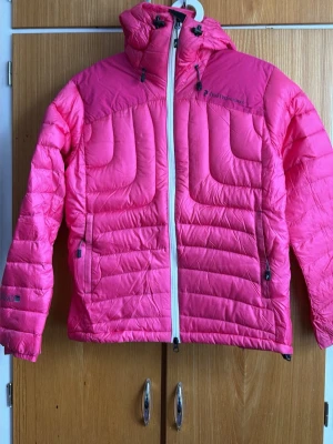 Rosa dunjacka Peak Performance - Snygg rosa dunjacka från Peak Performance med huva och vit dragkedja. Jackan har två sidofickor med dragkedja och justerbar huva med snörning. Quiltad design och lätt glansigt material som håller värmen under kalla dagar.