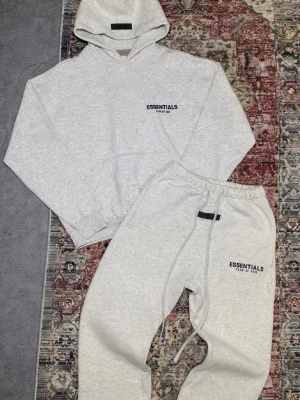 Grå Essentials hoodie & byxor set - Mysigt loungewear-set från Essentials med hoodie och matchande byxor i ljusgrå färg. Båda plaggen har svart Essentials-logga på bröst och lår. Hoodien har känguruficka och byxorna har dragsko i midjan. Perfekt för chill dagar hemma.