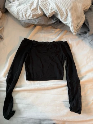 Svart mesh offshoulder topp - Trendig svart offshoulder topp i meshmaterial med långa ärmar. Toppen är croppad och har en tight passform som framhäver siluetten. Perfekt för dig som vill ha en edgy och stilren look.