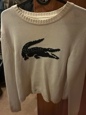 Vit stickad tröja från Lacoste - Säljer en vit stickad tröja från Lacoste med den klassiska svarta krokodilen på bröstet. Tröjan har rund halsringning och långa ärmar. Perfekt för dig som gillar stilrena plagg med ikoniska detaljer.