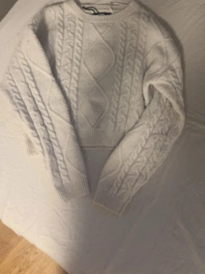 Vit kabelstickad tröja - Säljer en vit kabelstickad tröja med klassiskt flätmönster och ribbade muddar vid ärmslut och nederkant. Tröjan har rund halsringning och är lite croppad i modellen. Perfekt för dig som gillar en mysig och trendig look. Använt max 10 ggr för 1 år sen❤️