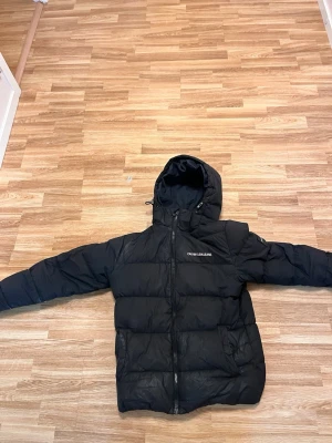 Calvin klein jacka - Säljer en svart pufferjacka från Calvin Klein Jeans med huva och dragkedja framtill. Jackan har logotypen tryckt på bröstet och en patch på ärmen. Perfekt för kalla vinterdagar och har en klassisk, stilren look. Den är en litet hål i en vänstra armen.