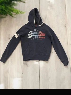 Mörkblå Superdry hoodie med tryck - Mörkblå hoodie från Superdry med stor logga och japansk text på bröstet. Klassisk känguruficka framtill, dragsko i huvan och tryck på ena ärmen. Perfekt för dig som gillar streetwear och vill ha en snygg och bekväm hoodie.
