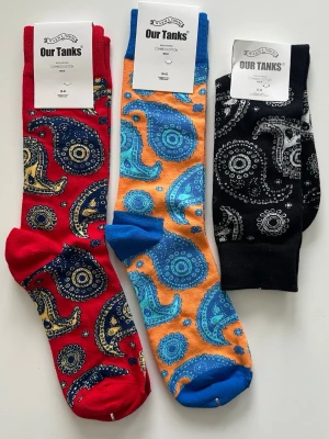3-pack NYA Färgglada paisleymönstrade strumpor - Tre par strumpor från Our Tanks i storlek 36-43 med paisleymönster i olika färger: ett par i rött och marinblått, ett par i orange och blått samt ett par i svart och vitt. Strumporna är i bomull och har en normal passform. Perfekta för dig som gillar färg och mönster. Helt NYA, så klart. 