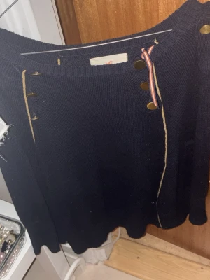 Svart stickad poncho med knappar - Säljer en svart stickad kortkjol med dekorativa guldknappar. Ponchon har en loose passform och är perfekt för dig som gillar en avslappnad men ändå trendig stil. Märket syns på etiketten men är otydligt på bilden.