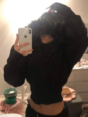 Svart pufferjacka med fuskpäls - Säljer en svart pufferjacka med stor huva och fuskpälsdetalj runt kanten. Jackan är midjekort och har en figurnära passform med dragkedja framtill. Perfekt för kalla vinterdagar och ger en snygg siluett.