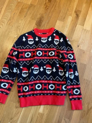 Mörkblå jultröja med tomtemotiv - Stickad jultröja i mörkblått med röda och vita detaljer. Tröjan har mönster med tomtar, granar och texten 'HO HO HO' runtom. Ribbstickade muddar och nederkant i rött. Perfekt för julens alla tillfällen!