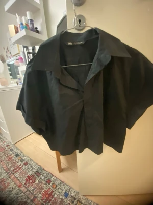 Svart croppad skjorta från Zara - Snygg svart croppad skjorta från Zara med bred krage och korta ärmar. Skjortan har en loose passform och är tillverkad i ett lätt tyg som känns skönt mot huden. Perfekt för dig som gillar en avslappnad men trendig stil.