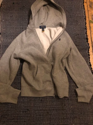 Grå hoodie från Polo Ralph Lauren - Klassisk grå hoodie från Polo Ralph Lauren med dragkedja framtill och liten mörkblå logga broderad på bröstet. Tillverkad i mjuk bomullsblandning och har en stor huva. Aldrig använd, nypris ligger på ca 1300 💓L i barnstorlek, cirka 13-16 år!