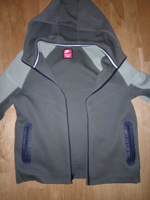 Nike tech fleece - Snygg hoodie från Nike i olivgrönt med ljusgrå detaljer på ärmarna. Dragkedja framtill, två fickor med mörka kantband och stor huva. Tillverkad i mjukt bomullsmaterial, perfekt för chill eller streetwear.