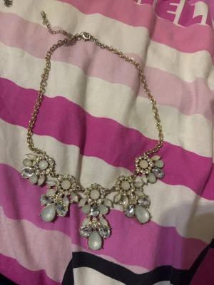 Guldigt statementhalsband med stenar - Säljer ett guldigt statementhalsband med stora vita och klara stenar i blomformade mönster. Halsbandet har en kedja i guldfärg och justerbar längd. Perfekt för dig som vill ha något som verkligen syns och ger en lyxig känsla till din outfit. 