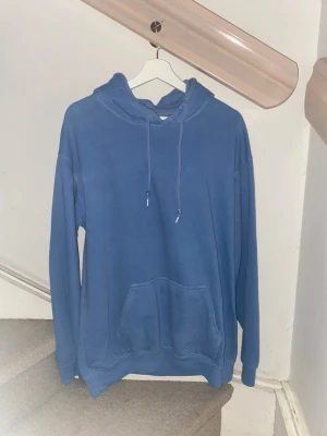 Blå hoodie med ficka framtill - En enkel blå hoodie med huva och dragsnören. Hoodien har en stor känguruficka framtill och ribbade muddar vid ärmslut och nederkant. Perfekt för chill dagar eller när du vill ha en avslappnad stil. Materialet känns mjukt och bekvämt. Storlek M