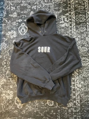 Svart Sour skateboards hoodie - Svart hoodie från SOUR med vit broderad text på bröstet. Klassisk modell med huva, känguruficka och ribbade muddar vid ärmar och nederkant. Perfekt för en avslappnad streetwear-look. Materialet ser ut att vara mjuk bomullsblandning. Tröjan har små vita färgfläckar på sig.