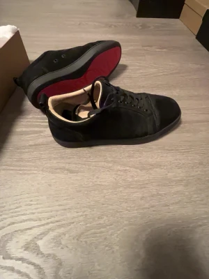 Svarta Louboutin sneakers med röd sula - Snygga svarta sneakers från Christian Louboutin med klassisk röd sula och lågt skaft. Ovandelen är i mocka och läder, med svarta skosnören och diskret design. Perfekta för dig som vill sticka ut med exklusiva detaljer och streetstyle-vibe.