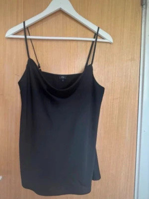 Svart linnetopp från River Island silk  - Svart linnetopp silk med tunna axelband och snygg draperad urringning. Enkel och stilren design som passar perfekt till jeans eller kjol. Tillverkad i ett mjukt och lätt material som känns skönt mot huden.