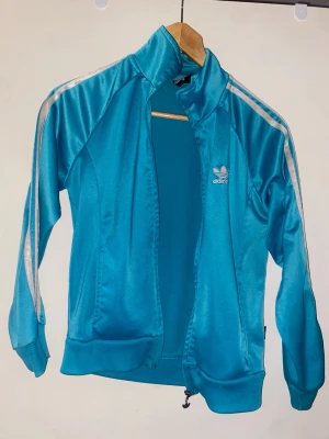 Turkos Adidas ziptröja med stripes - Snygg turkos ziptröja från Adidas med klassiska vita stripes längs ärmarna och broderad logga på bröstet. Tröjan har hög krage, muddar vid ärmslut och nederkant samt dragkedja framtill. Perfekt för en sportig och retro look.