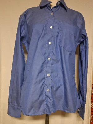 Blå klassisk skjorta med knappar - En stilren blå skjorta med lång ärm och klassisk krage. Skjortan har vita knappar framtill och en bröstficka på vänster sida. Perfekt för dig som gillar enkel och tidlös stil. Tillverkad i bomull för en skön känsla. Skjortan är aldrig använd. Men har varken förpackning eller prisetikett. Finns ej de förslaget på skick