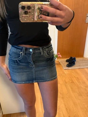 Mörkblå jeanskjol i kort modell - Säljer en snygg mörkblå jeanskjol i kort modell med klassiska fickor fram och bak samt knapp- och dragkedjestängning. Perfekt till vardags med en enkel topp eller hoodie. Tidlös denim-look som alltid är trendig.