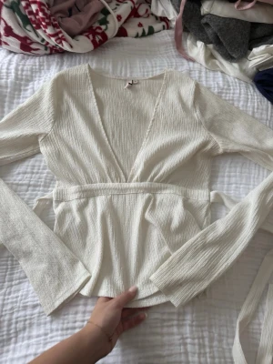 Vit omlottopp med knytband - Säljer en snygg vit omlottopp med långa ärmar och knytband i midjan. Toppen har en v-ringad skärning och är gjord i ett ribbat material som ger en cool struktur. Perfekt att styla med jeans eller kjol för en trendig look.