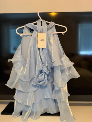 Ljusblå volangtop från YAS - Supersnygg OANVÄND  ljusblå halterneck-top från YAS i glansigt material. Toppen har flera lager volanger och knytning i halsen för en extra statement-look. Perfekt för dig som vill sticka ut med en unik och trendig topp. HELT NY