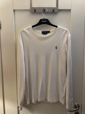 Vit långärmad tröja från Polo Ralph Lauren - Klassisk vit långärmad tröja från Polo Ralph Lauren med rund halsringning och den ikoniska broderade loggan på bröstet. Tillverkad i mjuk bomull som känns skön mot huden. Perfekt till jeans eller chinos för en clean och stilren look.