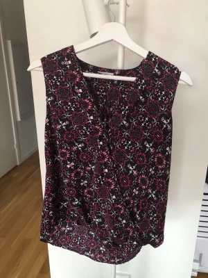 Mönstrad omlottopp i svart och rött - Säljer en gullig ärmlös omlottopp med svart bas och röda/lila samt vita detaljer i ett blommigt-liknande mönster. 