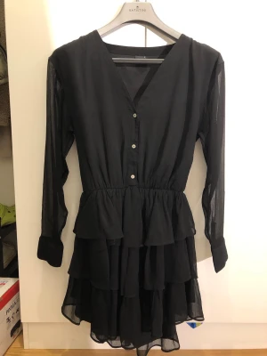 Svart volangklänning från Gina Tricot - Svart kortklänning från Gina Tricot med v-ringning, knappar framtill och långa ärmar. Klänningen har flera volanger i kjolen och är tillverkad i ett lätt, tunt tyg som ger ett luftigt intryck.