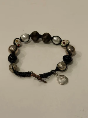 Pärlarmband med stjärna  - Handgjort armband med mixade pärlor i svart och silver, samt glitterpärlor med små röda stenar. Armbandet är av svart tråd och en silverfärgad berlock med stjärnmotiv. Har ett T-lås som har rostat 