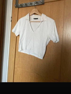 Vit kortärmad pikétröja från Zara - Säljer en vit, ribbad pikétröja från Zara med korta ärmar och krage. Tröjan har en djup v-ringning och är croppad i modellen, vilket ger en trendig och avslappnad look. 