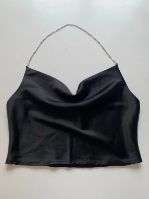 Svart satin bandeau topp med kedja - Snygg svart bandeau topp i satin med en unik kedja som axelband. Toppen har en draperad framsida och elastisk baksida för skön passform. Perfekt för dig som vill sticka ut med en trendig och elegant look.