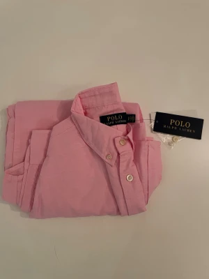 Ralph Lauren skjorta | Rosa - Säljer denna snygga rosa linneskjorta från Polo Ralph Lauren, med en ljusblå logga. Skjortan är helt ny då jag fick den i present för ungefär 1 år sedan. | Skick 10/10, Helt ny och märkeslapp + extra knappar medföljer. | Storlek: S | Pris kan diskuteras i pm! | MVH William