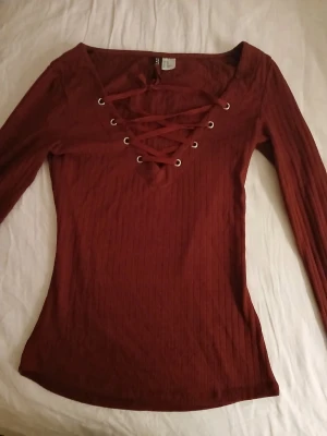 Vinröd ribbad topp med snörning - Snygg vinröd långärmad topp med ribbad struktur och snörning framtill i urringningen. Figurnära passform och djup v-ringning med metallöljetter. Perfekt för dig som vill ha en trendig och bekväm topp.