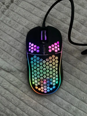 RGB gamingmus med honeycomb-design - Säljer en svart gamingmus med RGB-belysning och honeycomb-mönster på ovansidan. Musen har sladd och flera programmerbara knappar. Perfekt för dig som vill ha snygg belysning och lätt vikt när du spelar.