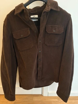 Brun manchester overshirt - Snygg brun overshirt i manchester från C med klassisk krage och två bröstfickor med knappar. Långärmad modell med knappstängning framtill, perfekt att slänga över en t-shirt för en chill look.