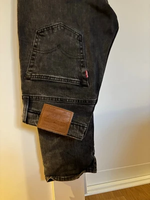 Svarta Levi's jeans med rak passform - Säljer ett par svarta jeans från Levi's med klassisk femficksdesign och rak passform. Jeansen har den ikoniska röda Levi's-lappen på bakfickan och patch i läder bak i midjan. Tillverkade i slitstark denim, perfekta till din vardagsstil.