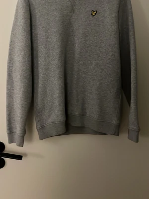 Grå sweatshirt från Lyle & Scott - Klassisk grå sweatshirt från Lyle & Scott med rund halsringning och ribbade muddar. Tröjan har den ikoniska gula loggan broderad på bröstet och är tillverkad i mjukt bomullsmaterial. Perfekt för en avslappnad och stilren look.
