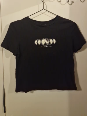 Svart croppad t-shirt med månfas-tryck - Svart croppad t-shirt med tryck av månens faser och texten 'to the moon & back' på bröstet. T-shirten har rund halsringning och korta ärmar. Perfekt för dig som gillar minimalistisk stil med en twist.