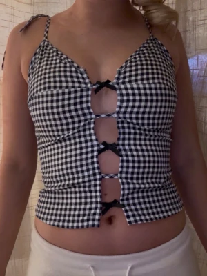 Rutig svartvit linnetopp med rosetter - Trendig linnetopp med svartvitt rutigt mönster och smala axelband. Toppen har flera små svarta rosetter framtill och öppna detaljer längs magen. Perfekt för dig som vill sticka ut med en unik och snygg topp.