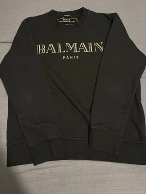 Svart sweatshirt från Balmain Paris - Säljer en svart sweatshirt från Balmain Paris med stort vitt tryck på bröstet. Tröjan har rund halsringning, långa ärmar och ribbade muddar vid ärmslut och nederkant. Perfekt för dig som gillar exklusiva streetwear-plagg.