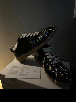 Maison Margiela sneakers med färgstänk - Unika svarta sneakers från Maison Margiela med färgstänk i vitt, gult, blått och rött. Skorna har vita snören och en klassisk gummisula. Perfekt för dig som vill sticka ut med en kreativ och konstnärlig vibe. Tillverkade i läder med låg profil.