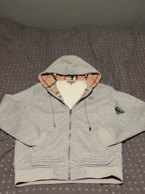 Burberry tröja - Storlek M/S!!!! Säljer en grå hoodie från Burberry med dragkedja och klassiskt rutigt foder i huvan. Hoodien har fickor framtill, justerbar dragsko i huvan och ribbade muddar. Perfekt för dig som gillar stilrena plagg med en twist av lyx. 