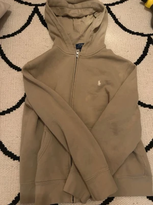Beige hoodie från Polo Ralph Lauren - Snygg beige hoodie från Polo Ralph Lauren med dragkedja framtill och klassisk huva. Tröjan har fickor på sidorna och den ikoniska vita logobrodyr på bröstet. Tillverkad i mjuk bomull för en skön sopig känsla.