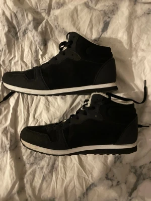 Svarta sneakers från Woolys med foder - Säljer ett par svarta sneakers från Woolys med vit sula och mjukt vitt foder på insidan. Skorna har klassisk snörning, högre skaft och är tillverkade i mockaliknande material. Perfekta för dig som vill ha både stil och komfort under kallare dagar.