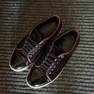 Lanvins - Snygga vinröda sneakers med svart lackad tå och svarta snören. Ovandelen är i mockaliknande material och sulan är vit. Skorna har metallöljetter och en stilren, låg profil som passar till många outfits.