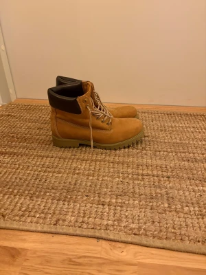 Bruna klassiska kängor med snörning - Säljer ett par bruna kängor i mockaimitation med grov sula och snörning. De har en vadderad svart kant runt ankeln för extra komfort och slitstarka detaljer. Perfekta för dig som vill ha en tidlös och robust look till hösten.