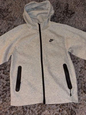 Nike tech fleece kofta grå  - Snygg ljusgrå hoodie från Nike med svart dragkedja framtill och svarta detaljer på fickorna. Klassisk Nike-logga på bröstet och huva för extra stilpoäng. Perfekt för chill dagar eller när du vill ha en sportig look.